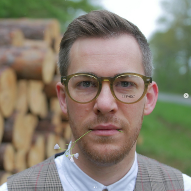Mann mit Brille und Blume im Mund, vor einem Holzstapel im Freien.