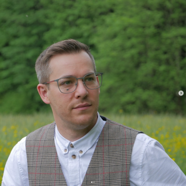 Mann mit Brille und Weste, steht in einer grünen Wiese mit Blumen.