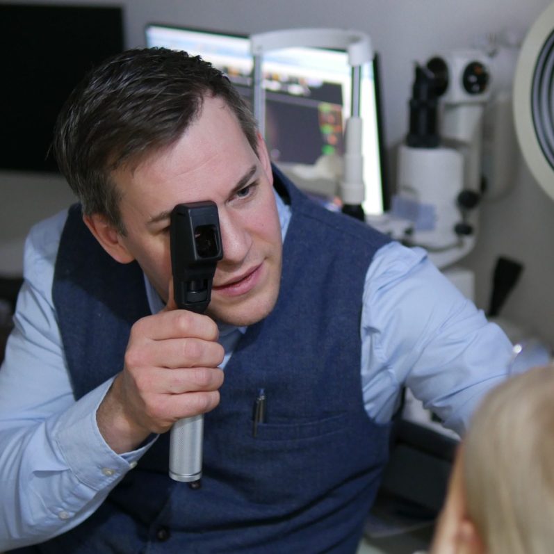 Herr Schötschel mit Skiaskop Optiker untersucht das Auge einer Patientin mit einem Skiaskop.