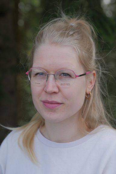 Junge Frau mit blonden Haaren, Brille und hellgrauem Oberteil, vor unscharfem Hintergrund.