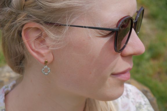 Porträt einer Frau mit Sonnenbrille und Ohrringen, im Freien vor grünem Hintergrund.
