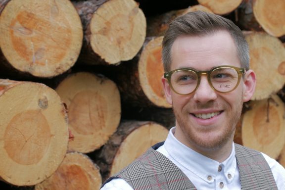 Junger Mann mit Brille lächelt vor einem Hintergrund aus Baumstämmen.
