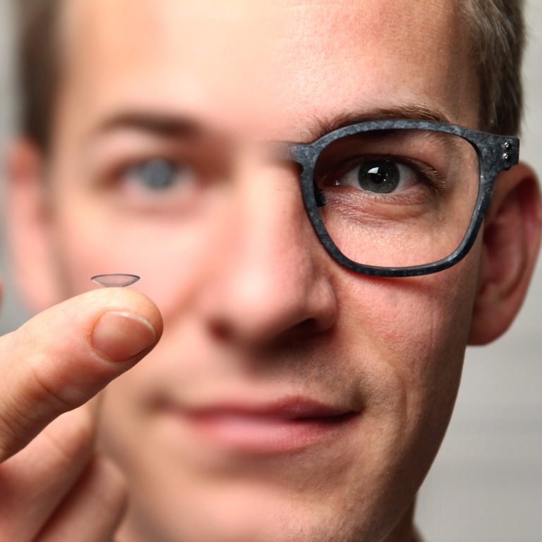 Orho-K Linsen Mann hält eine Kontaktlinse und blickt durch eine Brille mit nur einem Glas.
