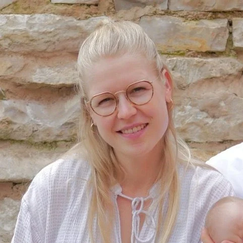 Bild von Anika Schötschel Frau mit blonden Haaren, Brille und weißem Oberteil, lächelt vor einer Steinwand.