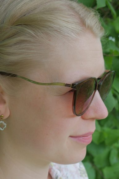 Frau mit blonden Haaren, Sonnenbrille und Ohrringen, vor grünem Hintergrund.