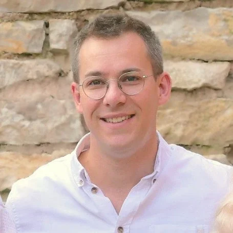 Bild von David Schötschel Mann mit Brille und kurzem, grauem Haar, lächelt vor einer Steinmauer.